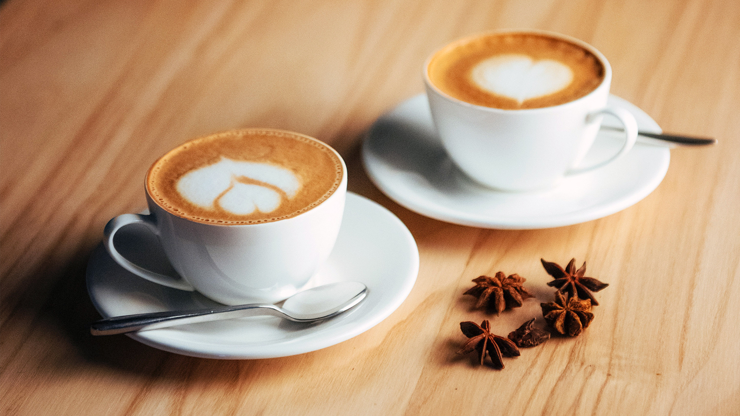 Na jasnym drewnianym blacie stoją dwie filiżanki cappucino a obok nich leżą gwiazdki anyżu.