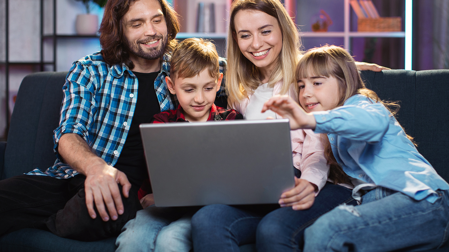 Rodzina siedzi na kanapie i przygląda się temu, co jest wyświetlane na ekranie laptopa.